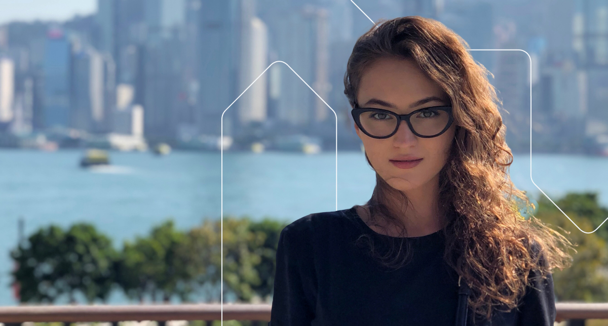 A woman stands in front of the Hong Kong skyline, she has long, brown hair and wears glasses